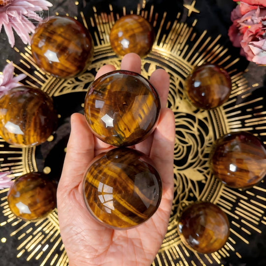Hand holding polished tiger eye crystal spheres for protection and spiritual growth, surrounded by decorative flowers and gold patterns.
