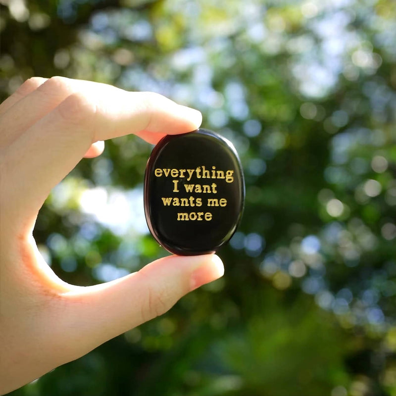 Hand holding black oval stone with gold quote 'everything I want wants me more' for spiritual growth