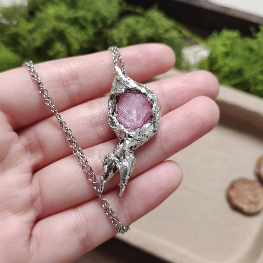 Handmade pink crystal pendant necklace with textured silver setting, held in hand against green moss background