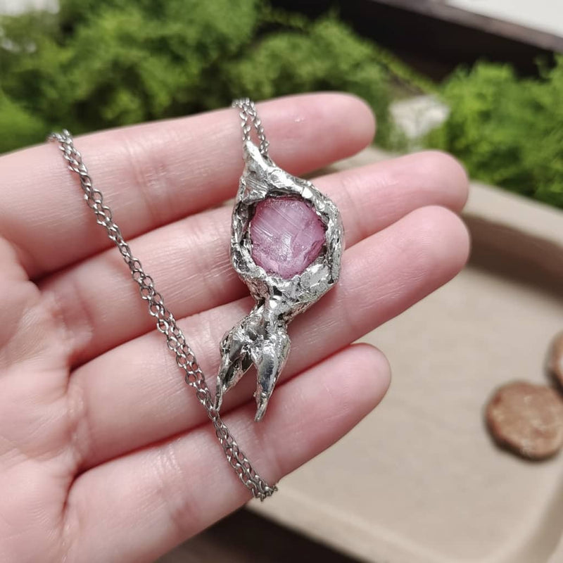 Handmade pink crystal pendant necklace with textured silver setting, held in hand against green moss background