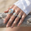 Hand wearing handmade seashell and aquamarine gemstone rings on driftwood, beach-inspired spiritual jewelry