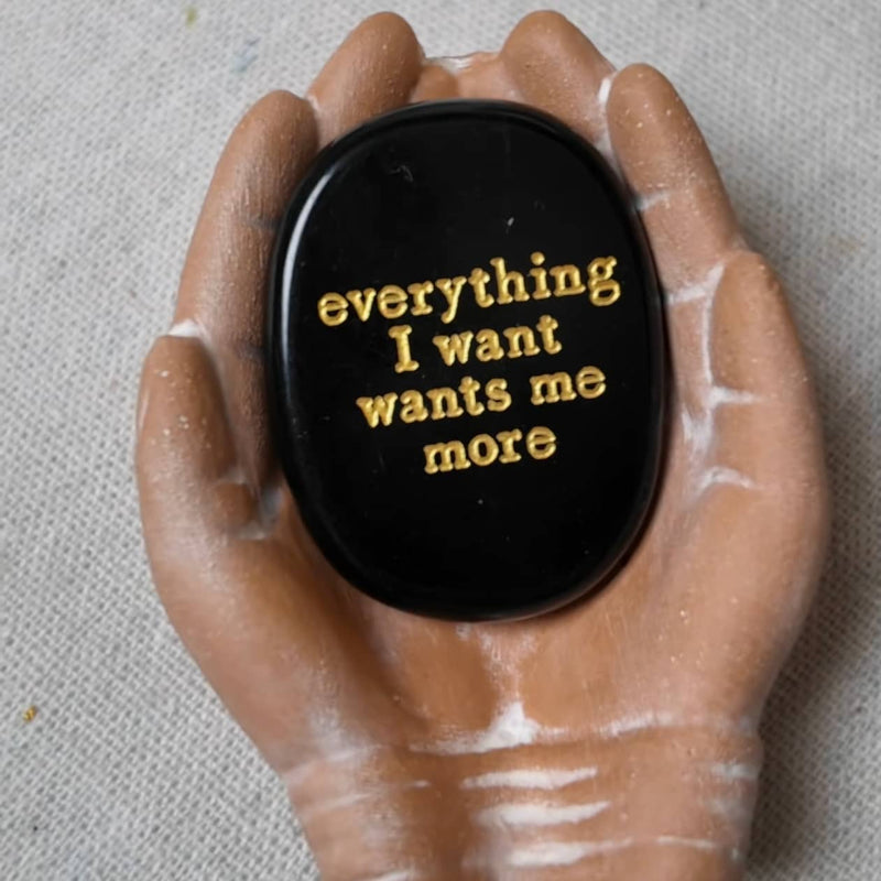 Black oval stone with gold text 'everything I want wants me more' held in hands, spiritual affirmation crystal