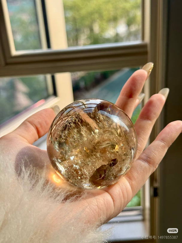 Hand holding a clear crystal sphere with golden inclusions, ideal for healing and spiritual growth near a sunlit window.