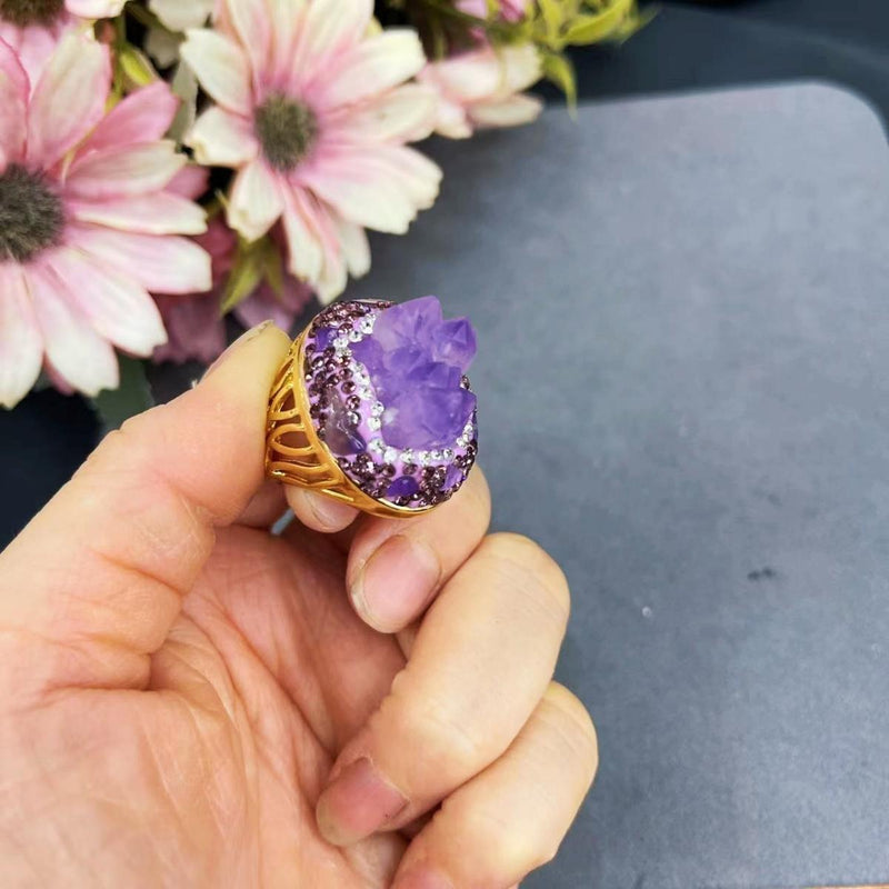 Amethyst crystal ring with gold band and gemstone accents, held near pink flowers for spiritual jewelry styling