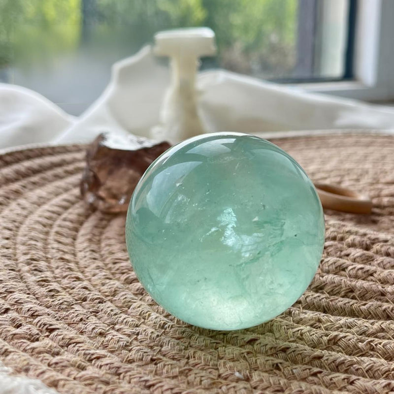 Smooth green fluorite sphere for spiritual healing and chakra balance, displayed on woven mat near window