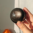 Hand holding a polished black crystal sphere with shimmering inclusions, ideal for spiritual healing and chakra balance.