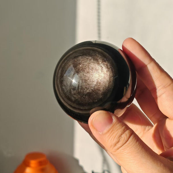 Hand holding a polished black crystal sphere with shimmering inclusions, ideal for spiritual healing and chakra balance.