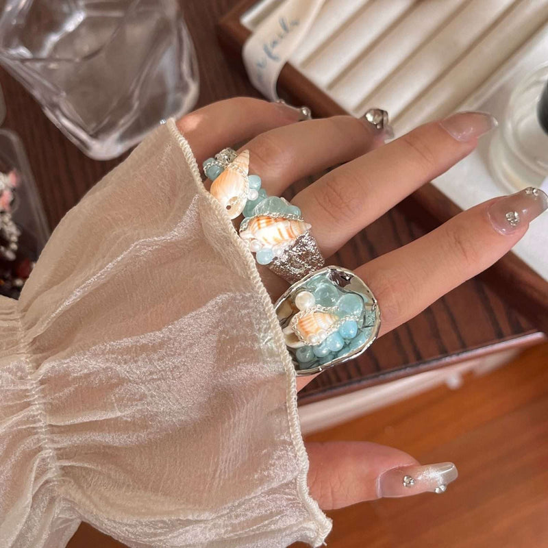 Hand wearing ocean-themed rings with seashells and blue beads, perfect for beach lovers and crystal jewelry fans.