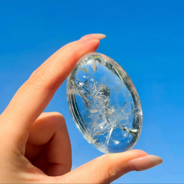 Clear quartz crystal egg with etched design, held against blue sky for spiritual healing and energy work