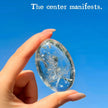Hand holding clear crystal with 'The center manifests' engraving against blue sky, spiritual healing stone