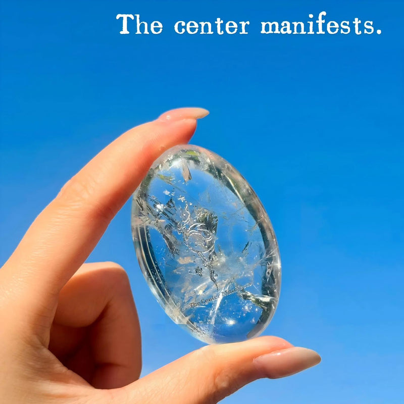 Hand holding clear crystal with 'The center manifests' engraving against blue sky, spiritual healing stone